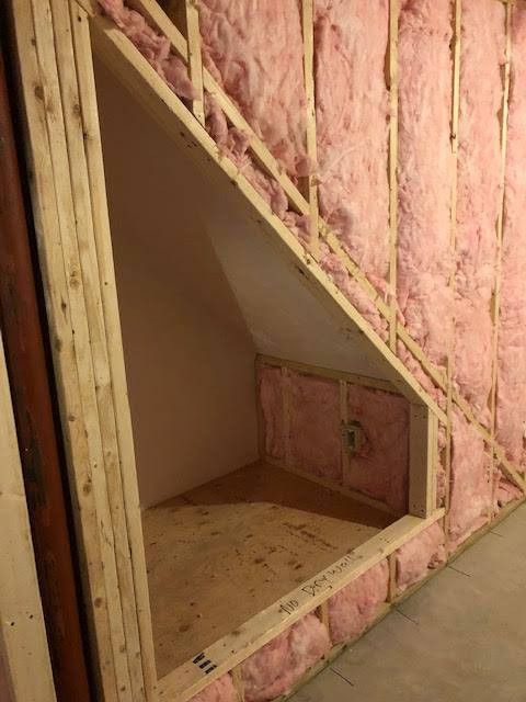 Framed, insulated storage space under stairs; unfinished, with bare wood and pink insulation.