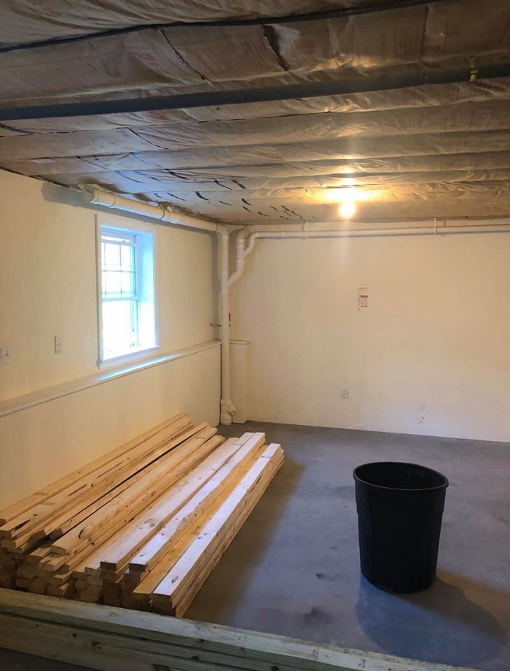 Basement with lumber, a window, and a black bucket. Beige walls and ceiling insulation.