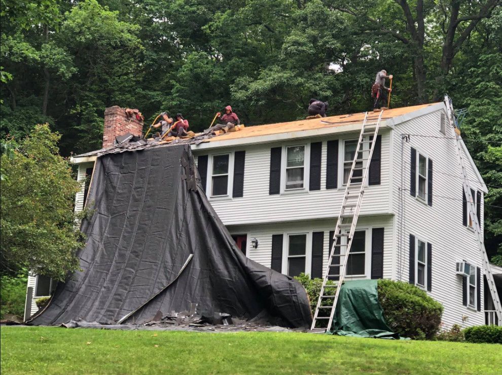 Roofers removing old shingles from a white house, with a black tarp covering part of the roof.
