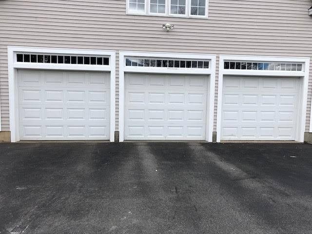 Three white garage doors with decorative window trim above, on a light-colored building, dark asphalt driveway.