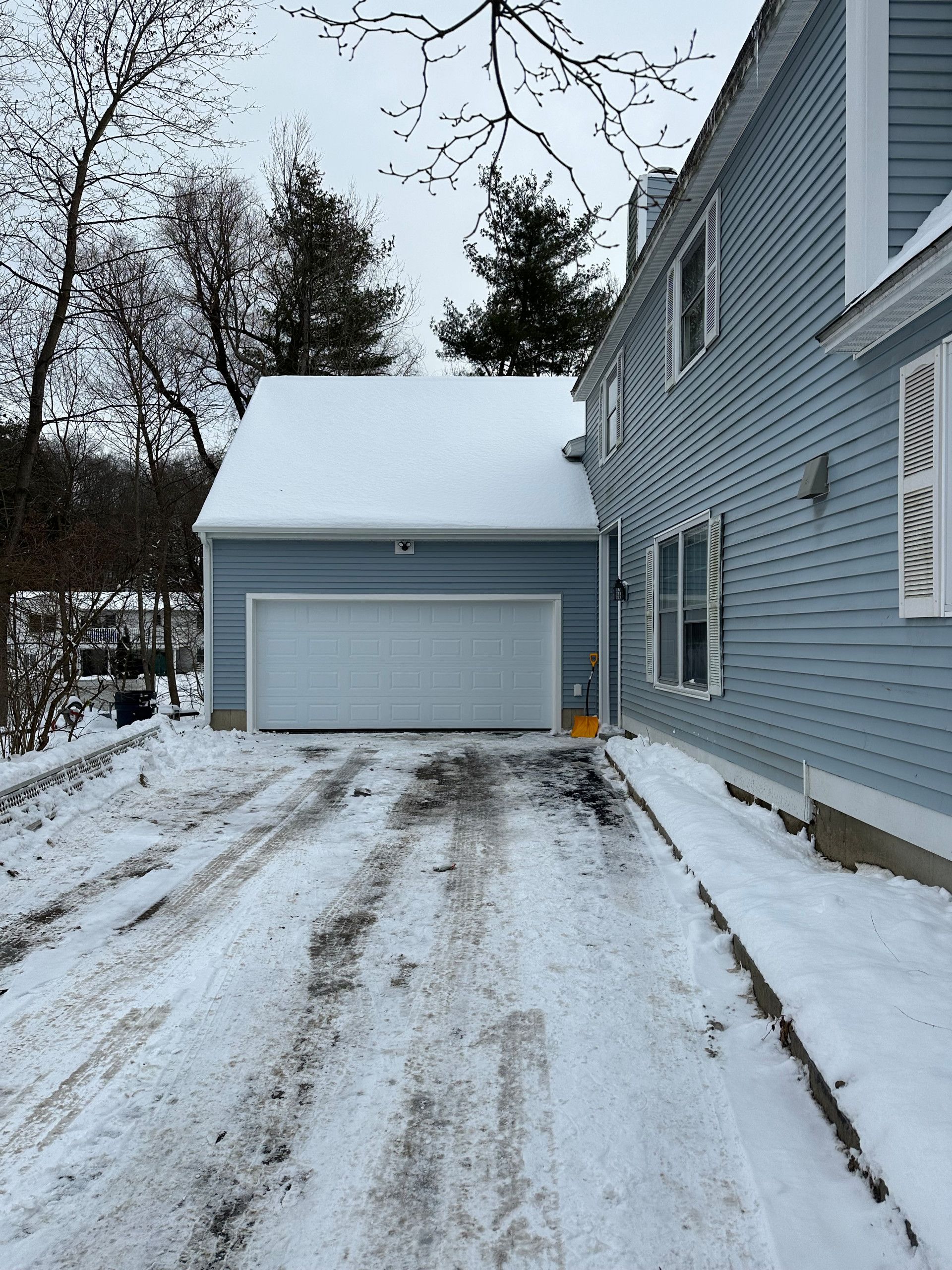 Snowy driveway leading to a light blue garage next to a blue house. Trees in background.