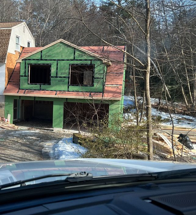 Two-story house under construction, green siding, red roof, garage doors open, snow on ground, woods in background.