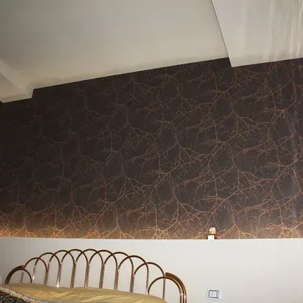 Parete scura con motivo ramificato color rame, sopra un divano dorato e una parete bianca.