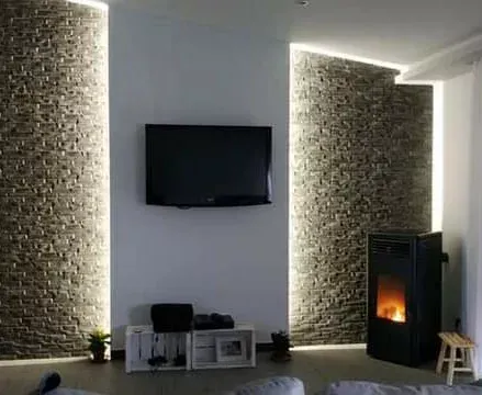 Soggiorno con TV a parete, pareti con elementi in pietra e illuminazione a incasso. Sono visibili anche un camino e un ripostiglio per la legna.