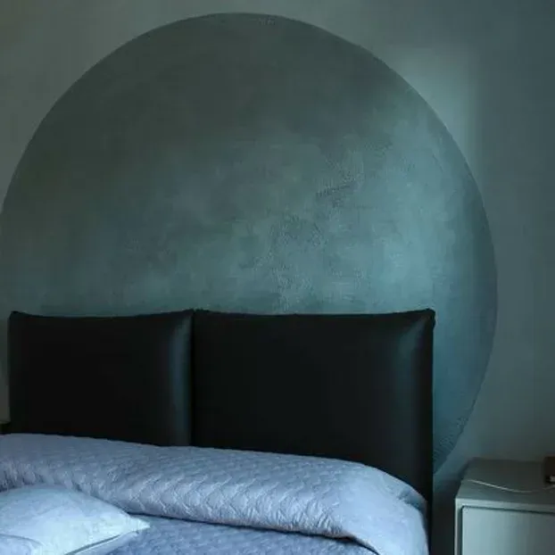 Testiera nera contro un cerchio dipinto di verde acqua su una parete della camera da letto; letto con lenzuola blu e un cuscino bianco.
