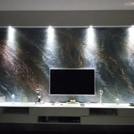 TV montata su una parete marmorizzata, illuminata da faretti, sopra un mobile bianco con oggetti decorativi.