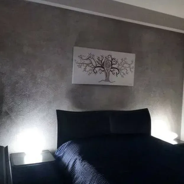 Camera da letto buia con un letto e un dipinto raffigurante un albero sulla parete. Le lampade laterali forniscono illuminazione.