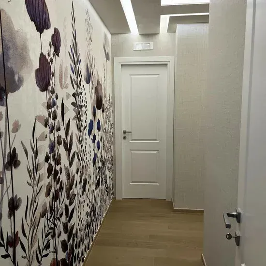 Corridoio stretto con carta da parati floreale sulla sinistra, porta bianca in fondo e pavimento in legno chiaro.