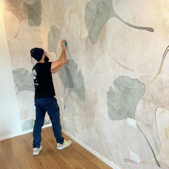 Uomo che installa carta da parati con un motivo a foglie grandi su una parete in una stanza con pavimento in legno.