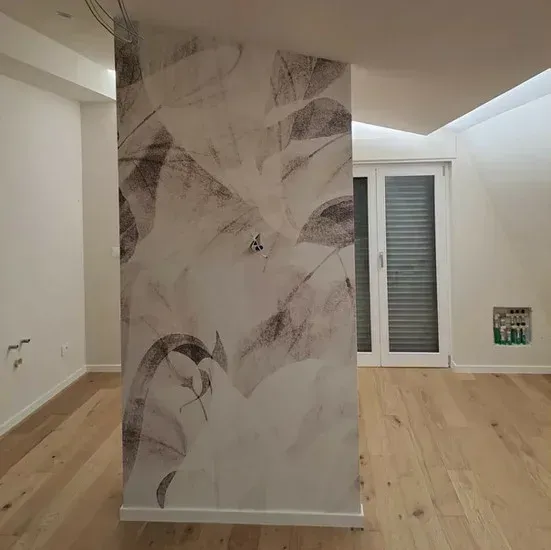 Interno con carta da parati floreale su una colonna centrale, pavimento in legno e pareti bianche.