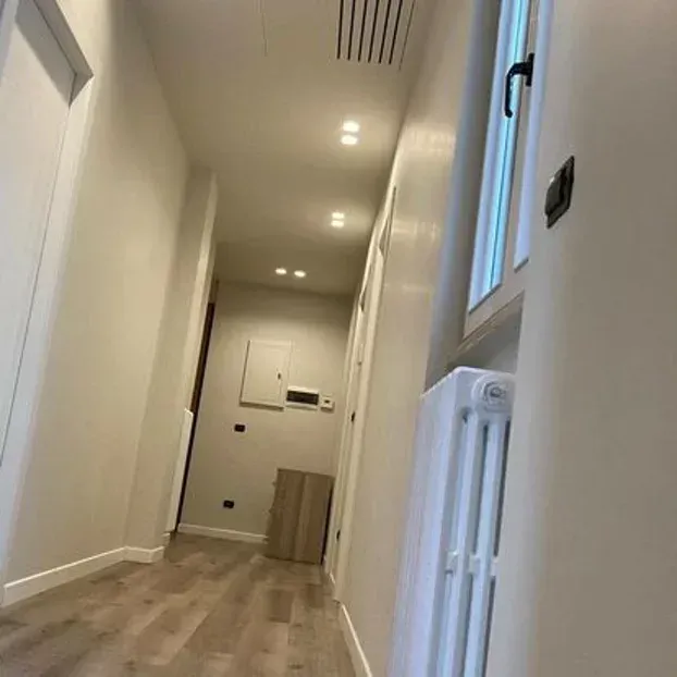 Corridoio stretto con pareti chiare, faretti a soffitto e pavimento in legno. Una finestra e un termosifone si trovano sulla destra.