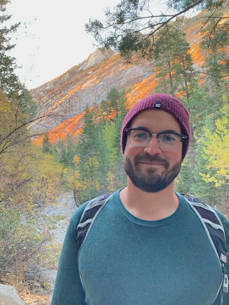 Man wearing a maroon beanie and glasses standing on a forest trail with sunlit mountains behind him
