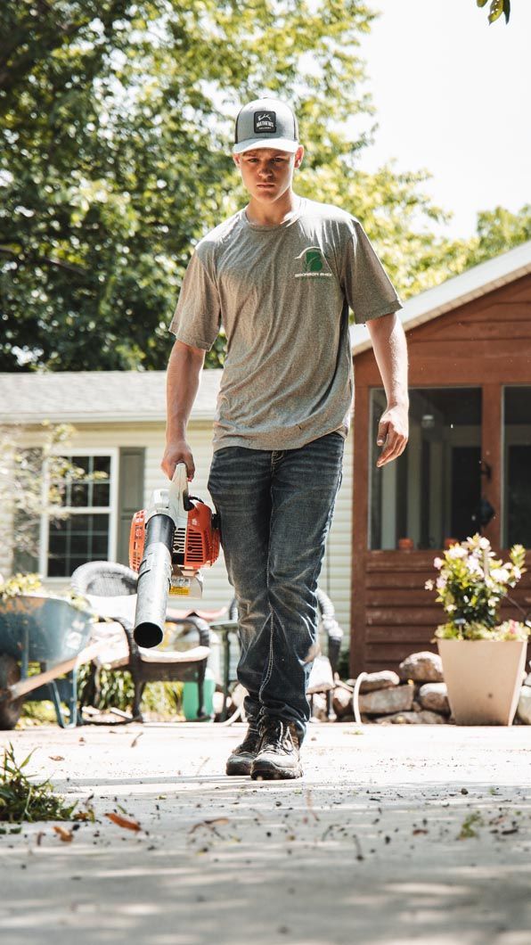 Man in jeans and t-shirt holds a leaf blower on a sunny patio near houses.