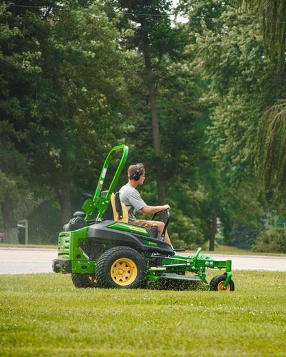 Man on a green and yellow John Deere zero-turn mower mowing grass in a grassy area with trees.