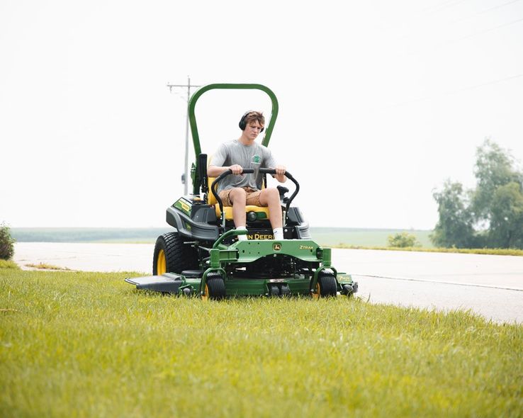 Man riding a John Deere mower, cutting grass near a road.