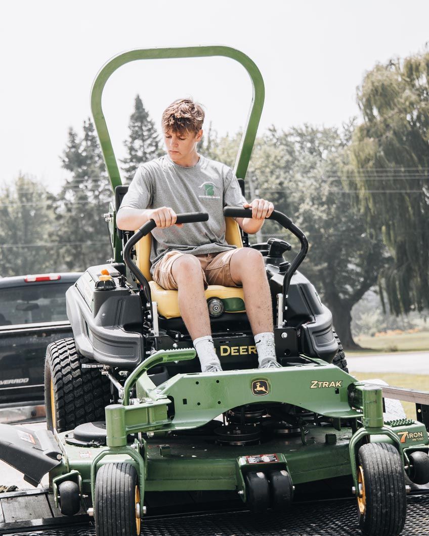 Teenager on a green and yellow John Deere zero-turn mower, wearing a t-shirt, sitting outside.
