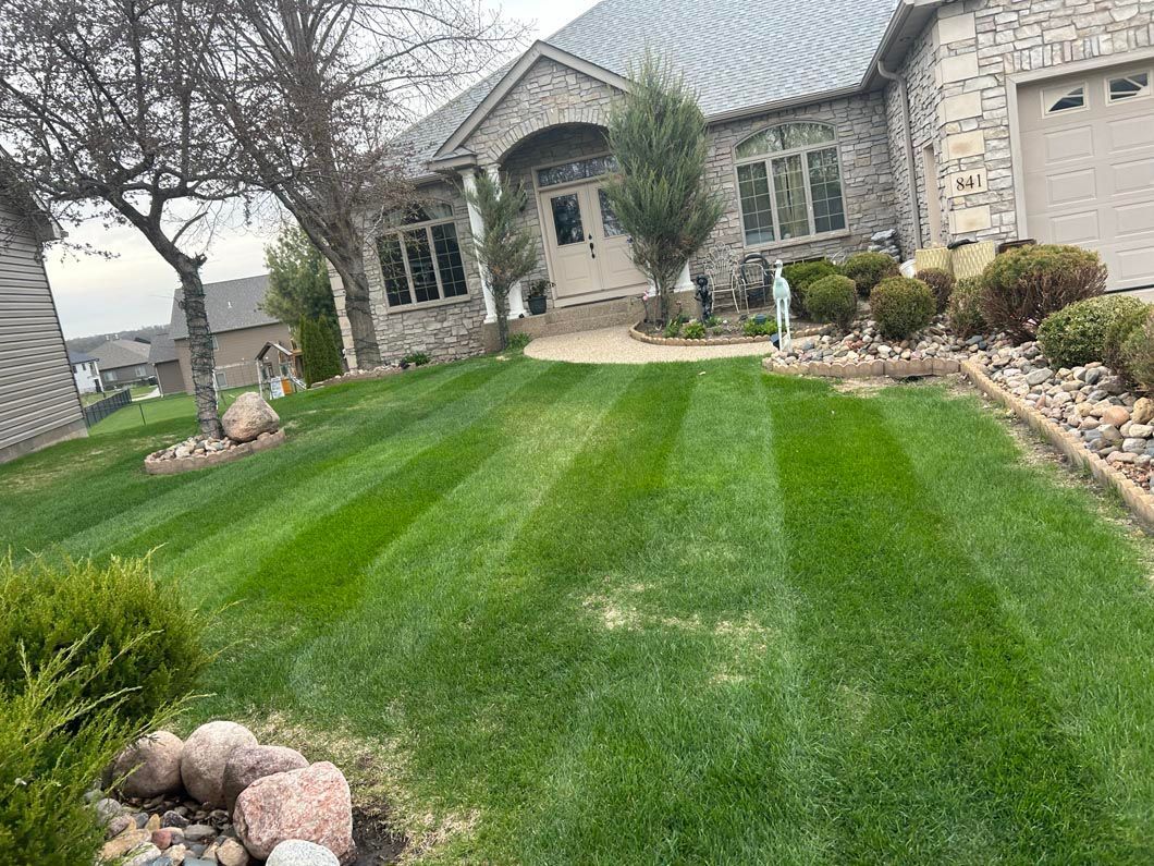 Well-manicured green lawn with striping, leading to a stone house with a driveway.