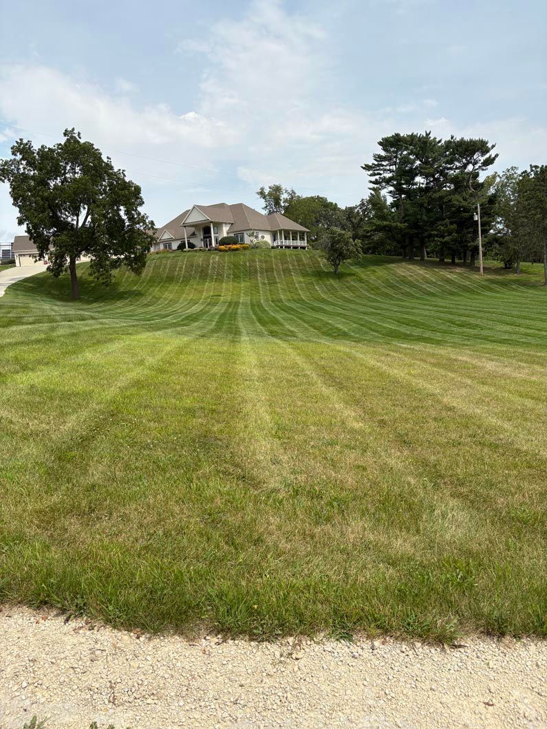 Lawn mowed in stripes leading uphill to a large house on a grassy hill under a cloudy sky.