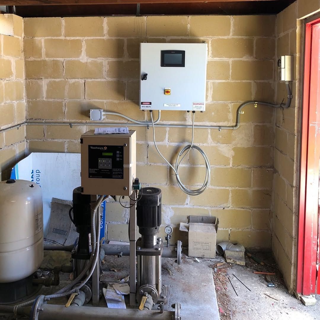 Water pump station with two pumps, control boxes, and a white tank against a beige block wall.