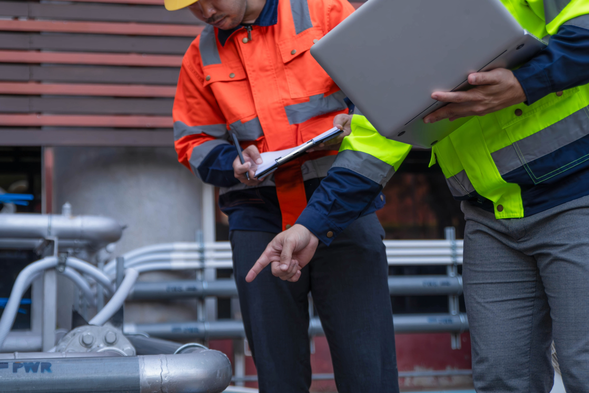 Two workers in safety vests and hard hats inspecting pipes, one pointing while the other holds a laptop and clipboard.