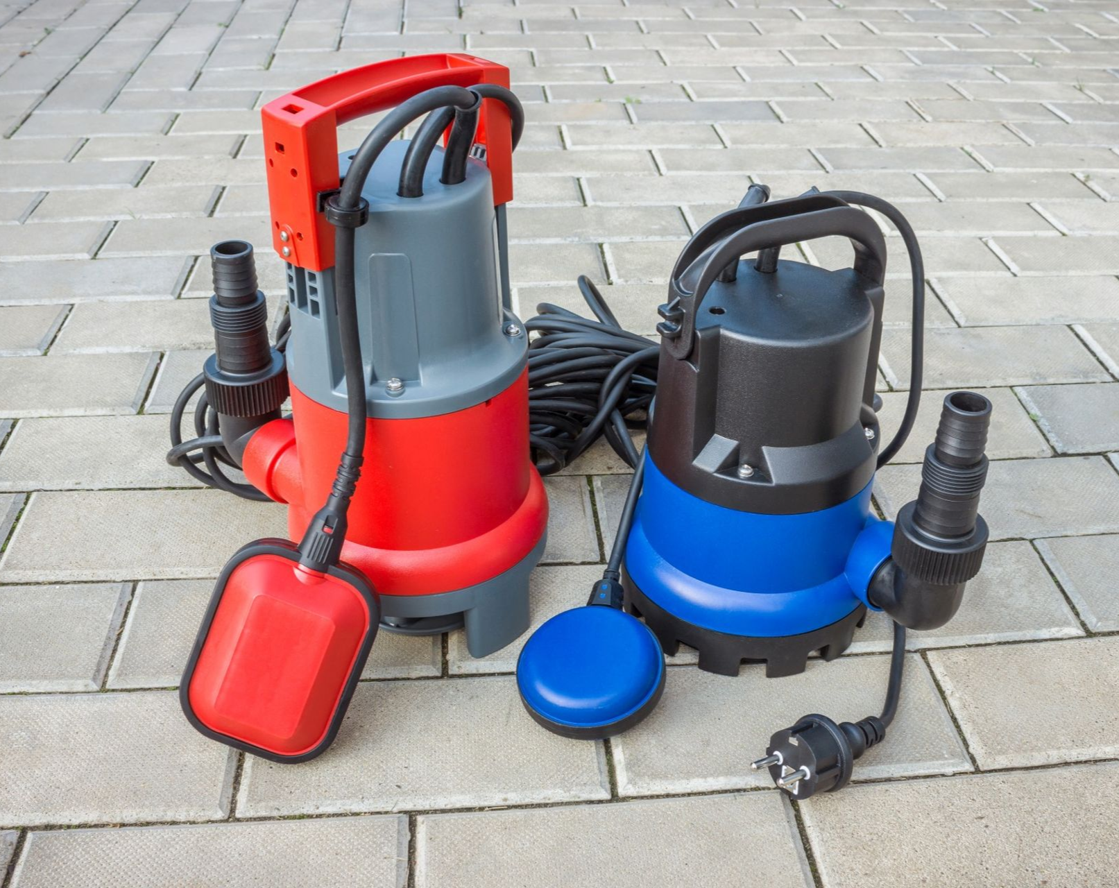 Two submersible water pumps, red and blue, on a brick patio. Each has a handle, hose connector, and float switch.