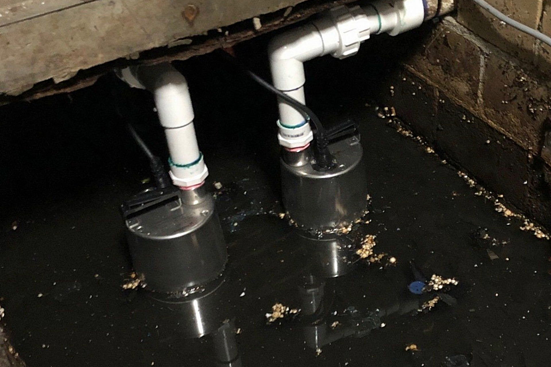 Two submerged sump pumps in a dark, flooded pit with white pipes and electrical cords.