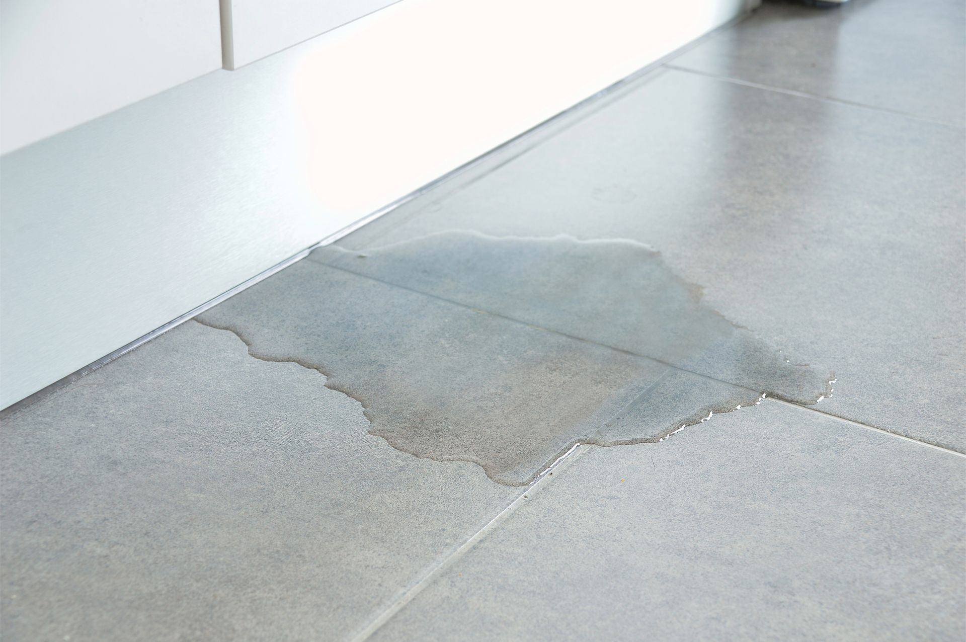 Water puddle on gray tiled floor next to a white baseboard.