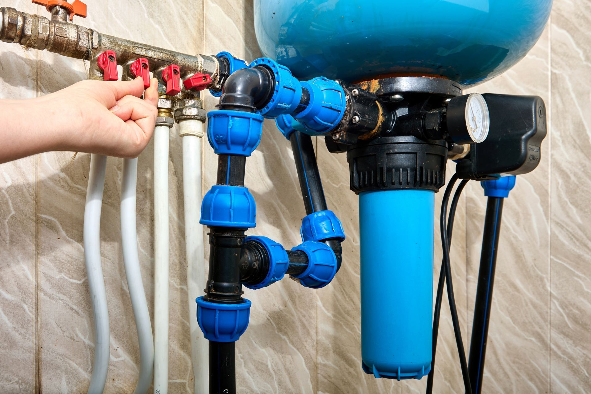 Hand turning a valve on water filtration system with blue and black pipes against a tiled wall.