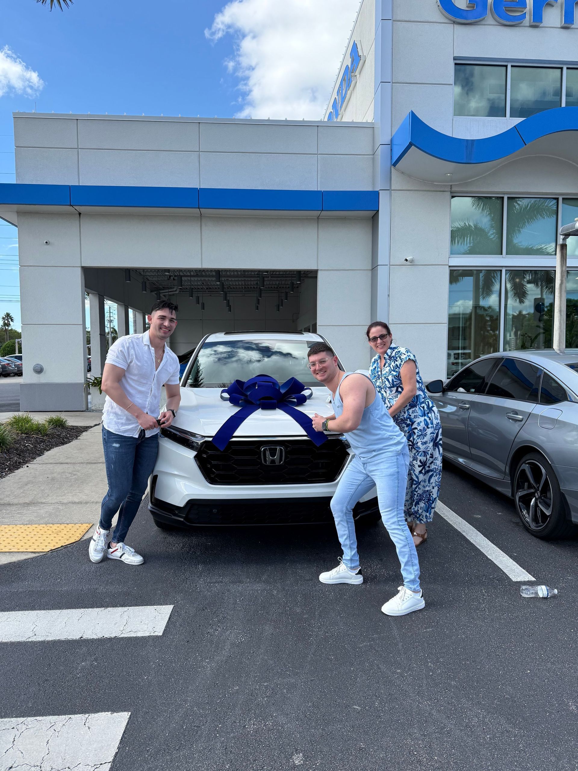 Three people pose with a new white Honda SUV in front of a dealership with a large blue bow.