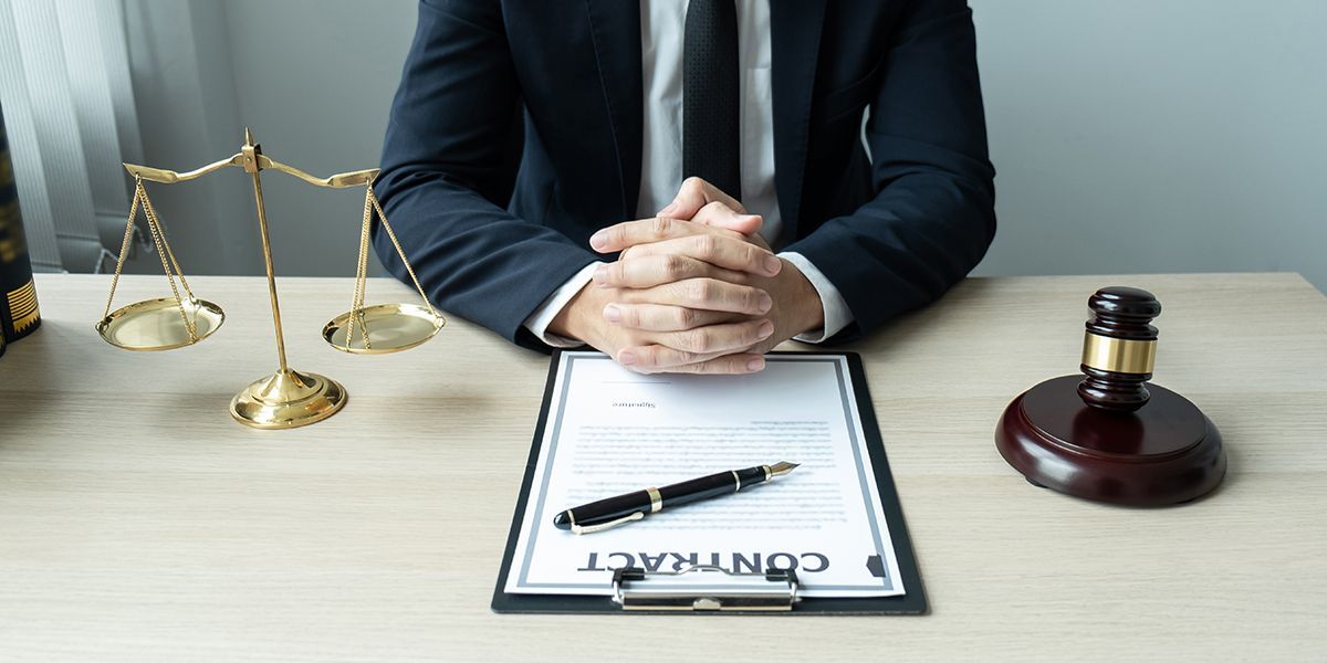 A person in a suit sits at a desk with scales, a gavel, and a contract.