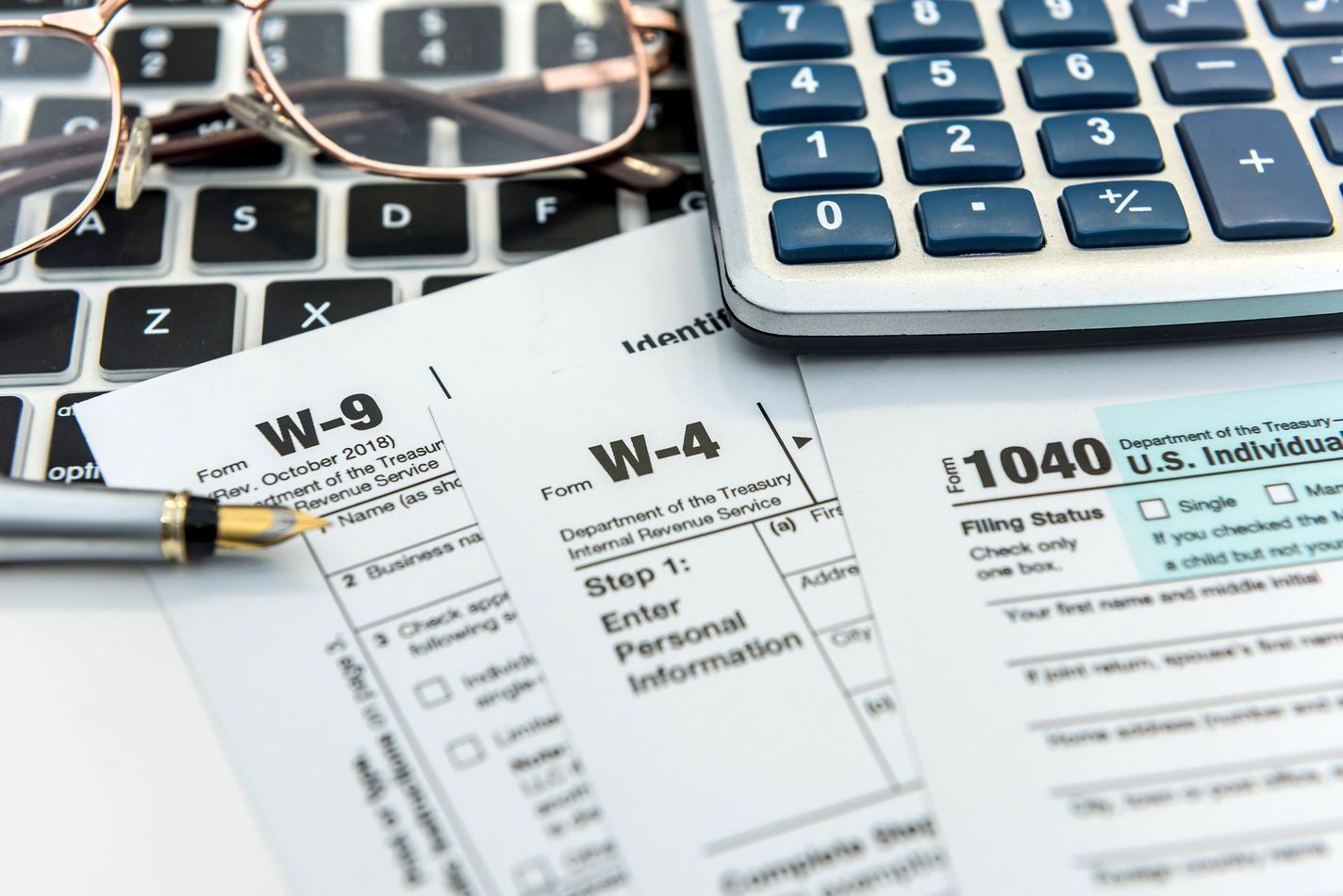 Tax forms, calculator, glasses, and pen on a laptop keyboard.
