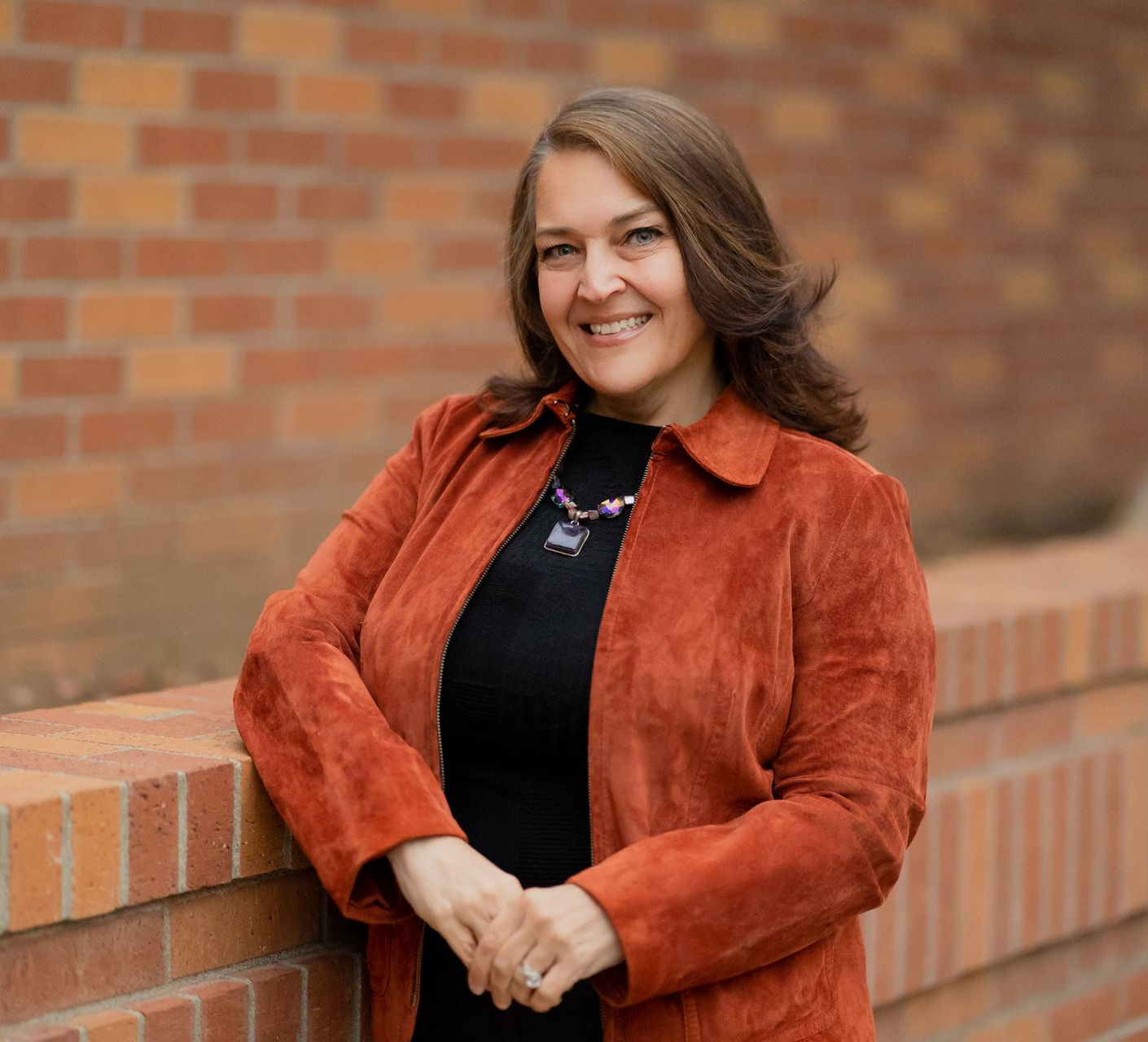Woman in orange jacket leans on a brick wall, smiling.
