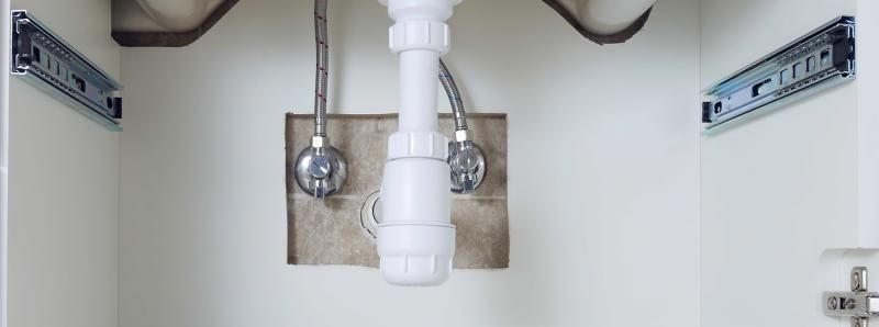 Under-sink cabinet interior with white walls, exposed chrome plumbing valves, a white drain pipe, and metal drawer slides.
