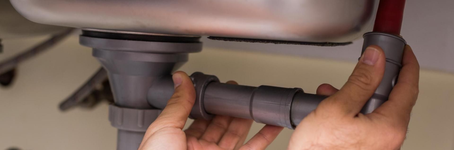 A person’s hands fitting gray plastic pipes together under a kitchen sink.