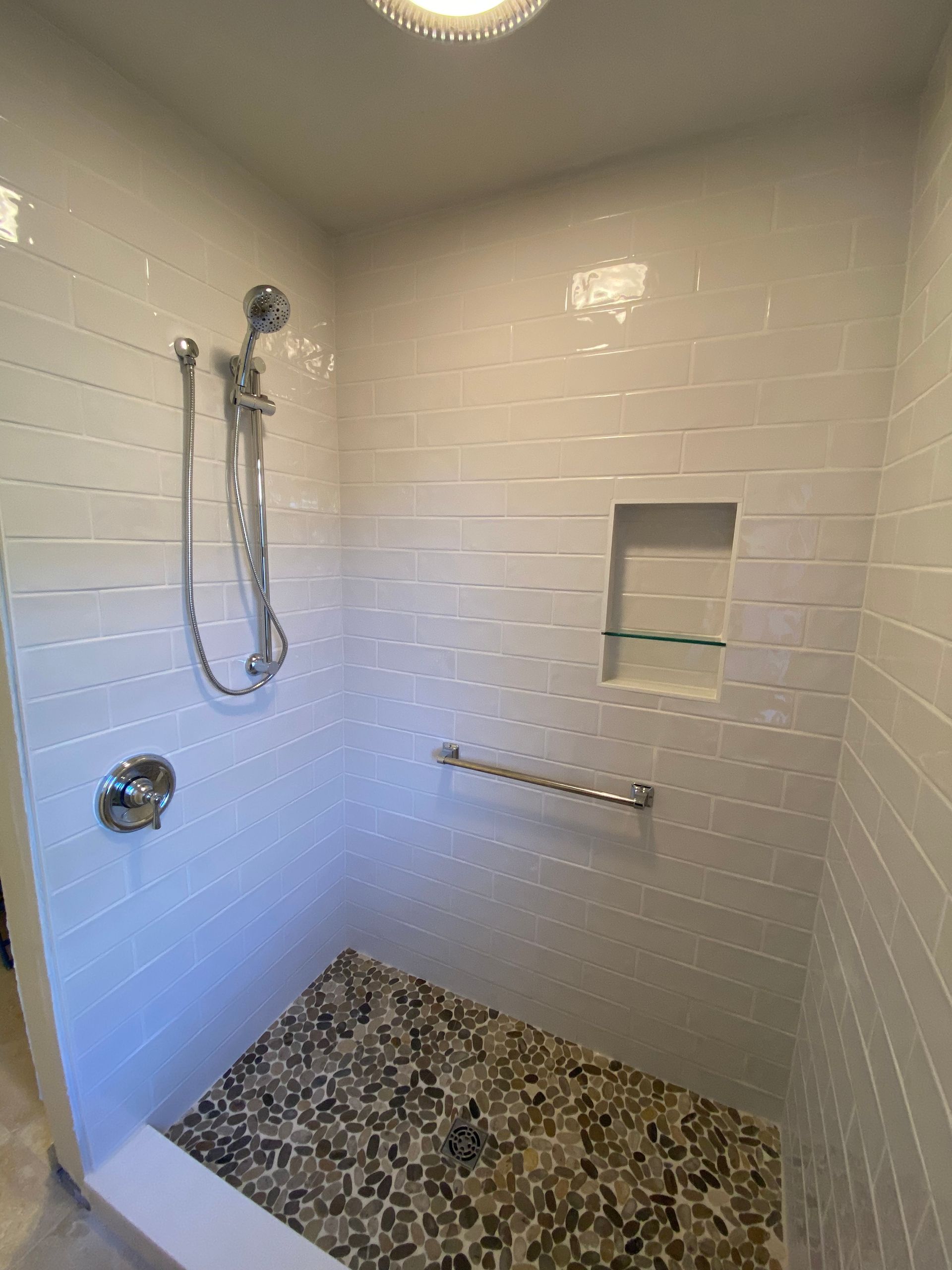 Bathroom shower with pebble floor, white tiled walls, and grab bar.