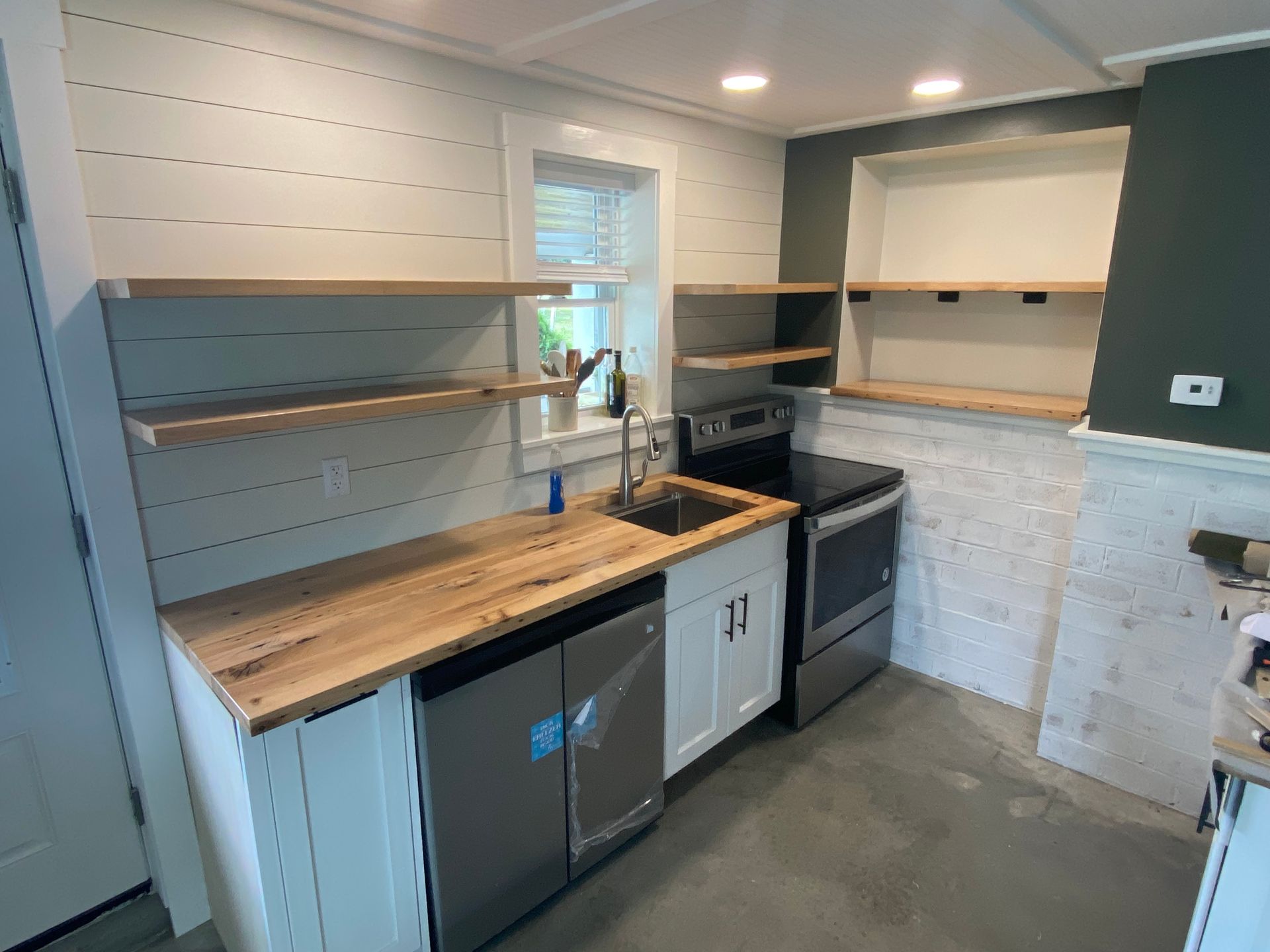 Small kitchen with white cabinets, wood countertops and shelves, stainless steel appliances, and a gray concrete floor.