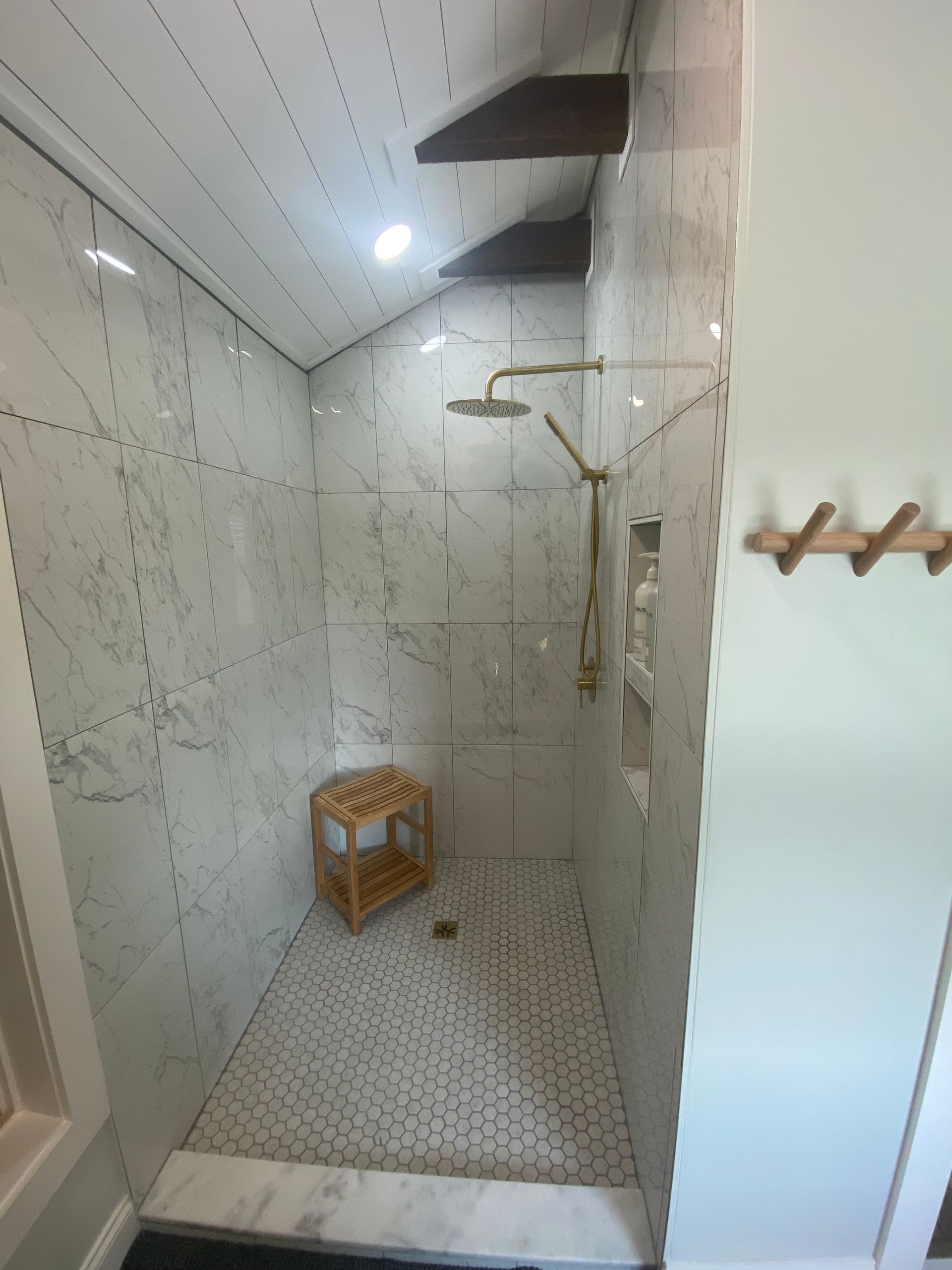 Bathroom with marble tiled shower, gold fixtures, wood bench, and textured floor.