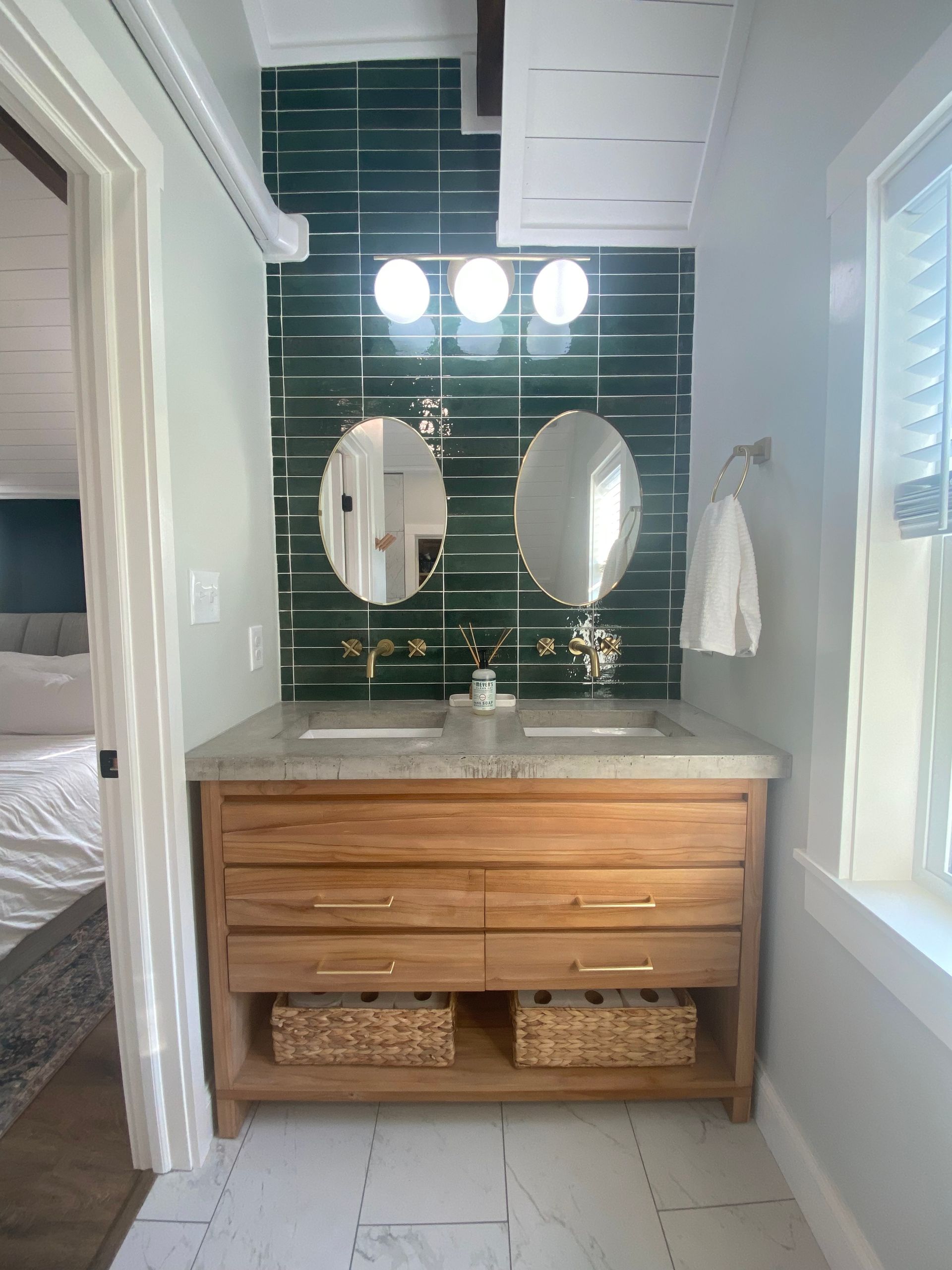 Bathroom with wooden vanity, green tile backsplash, two oval mirrors, and a window.