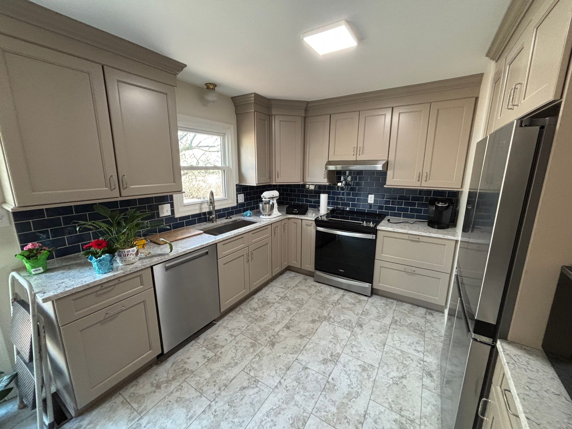 Tan kitchen cabinets, stainless steel appliances, and blue tiled backsplash. Gray floor.