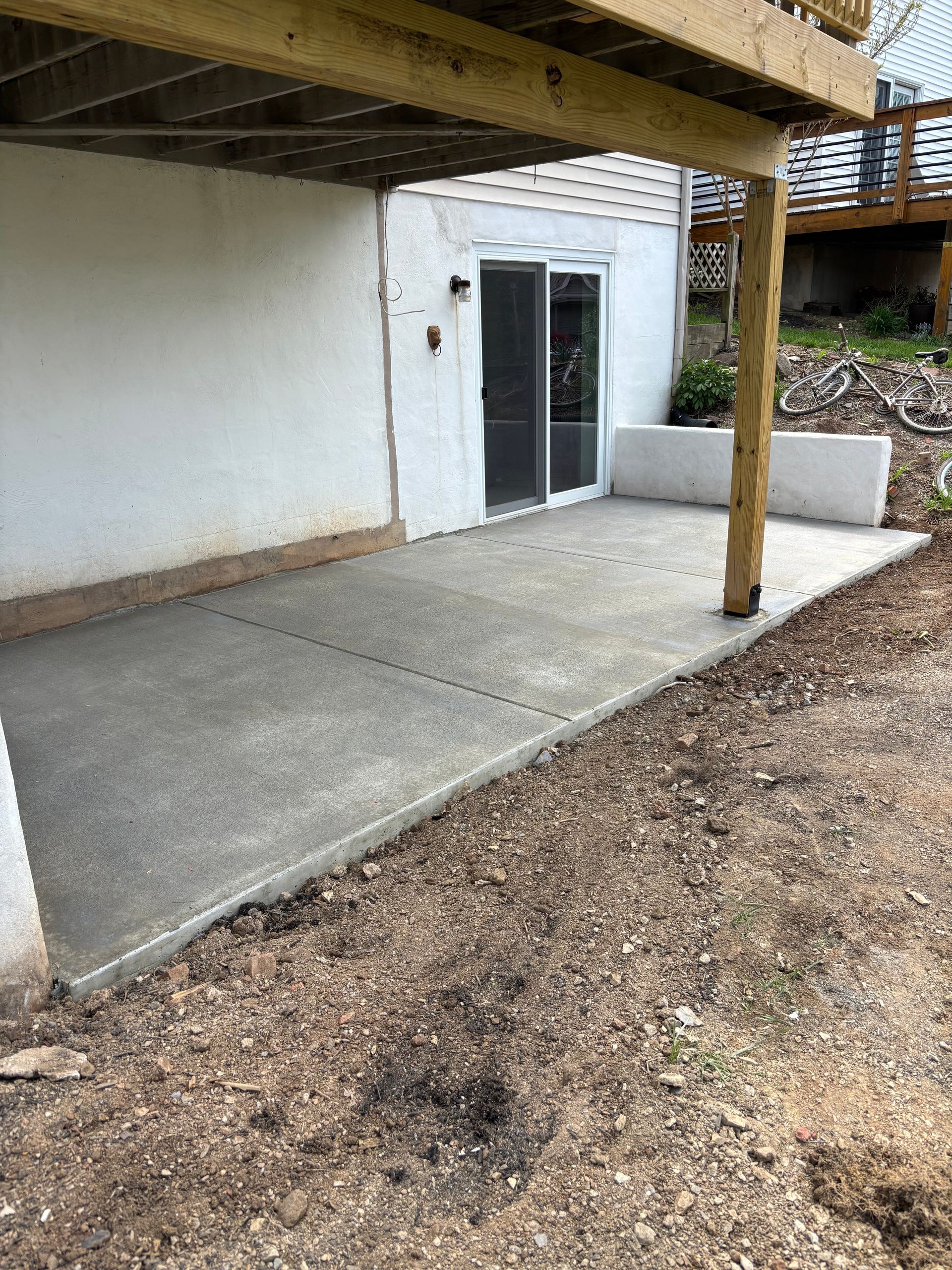 Concrete patio under a deck, next to a white wall with sliding glass door.