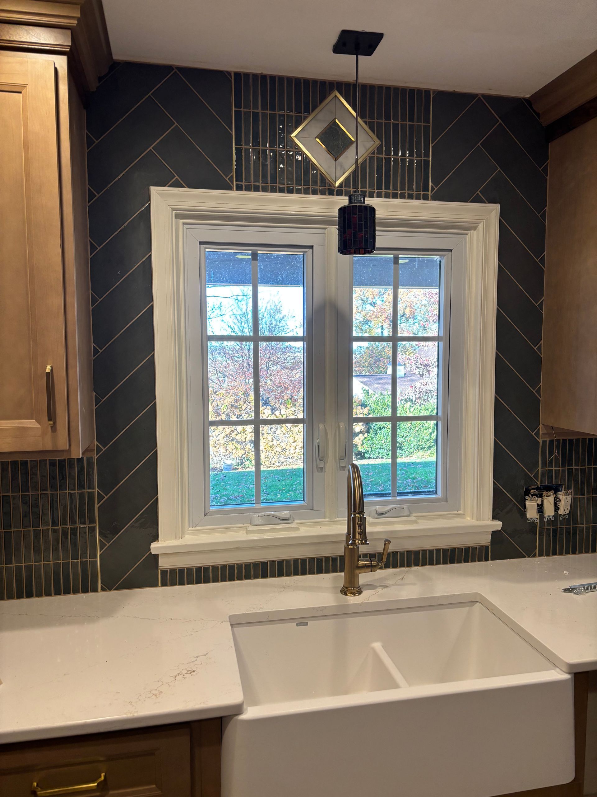 Kitchen sink with window above, herringbone tile backsplash, gold faucet, and decorative light fixture.
