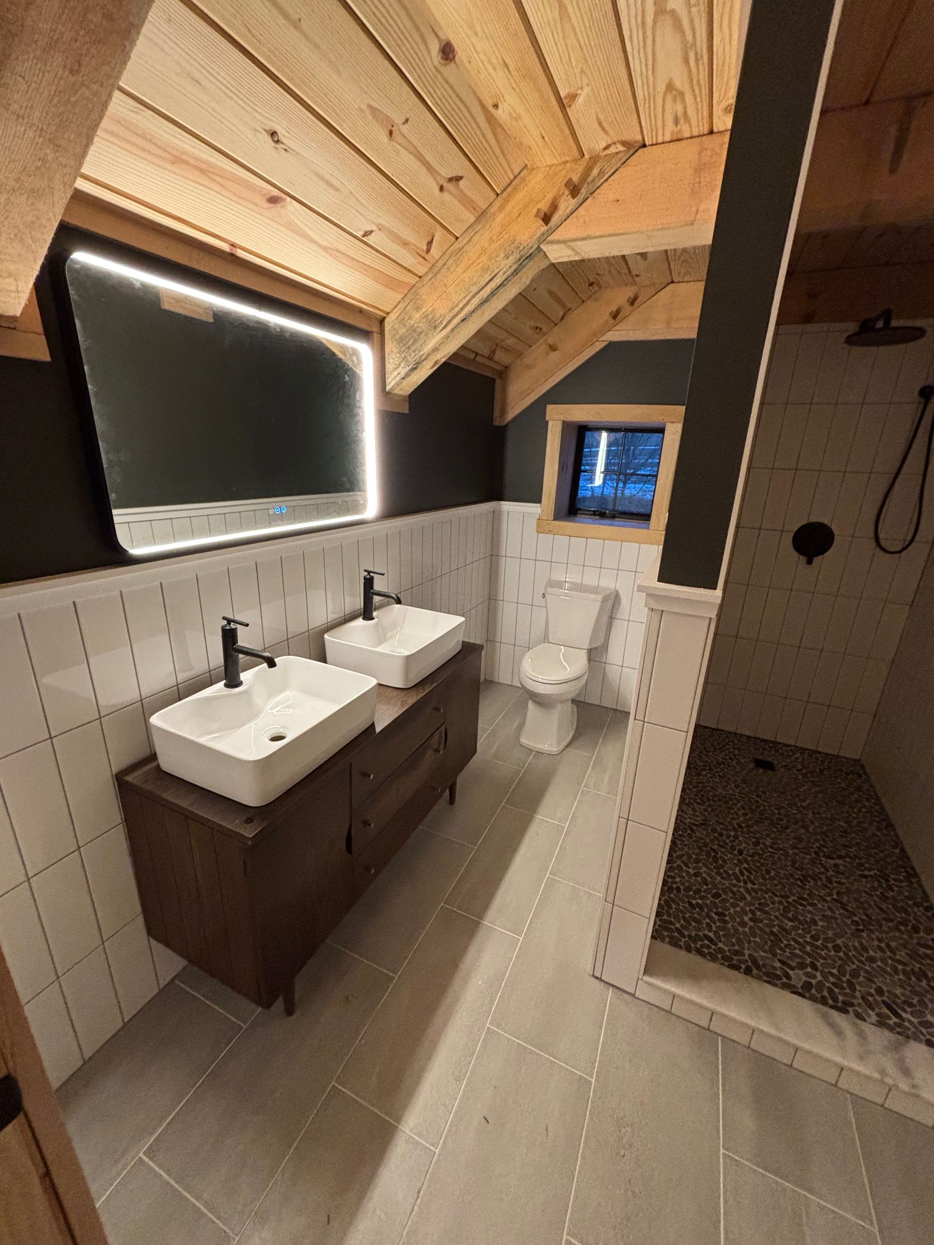 Bathroom with wood ceiling, dark walls, double sinks, a toilet, and a pebble-floored shower.
