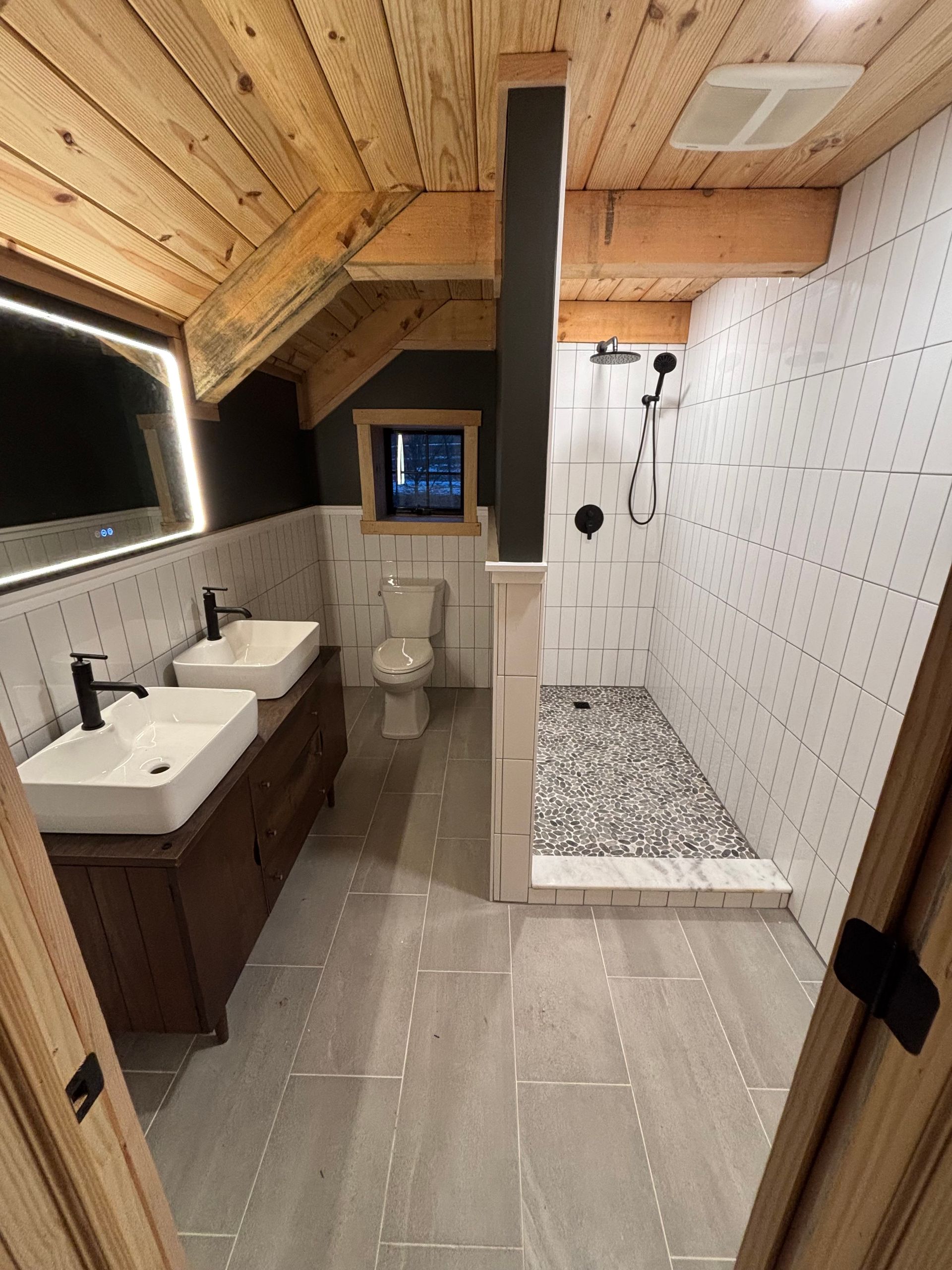Bathroom with wood ceiling, gray tile floor, white sinks, and a pebble-floored shower.