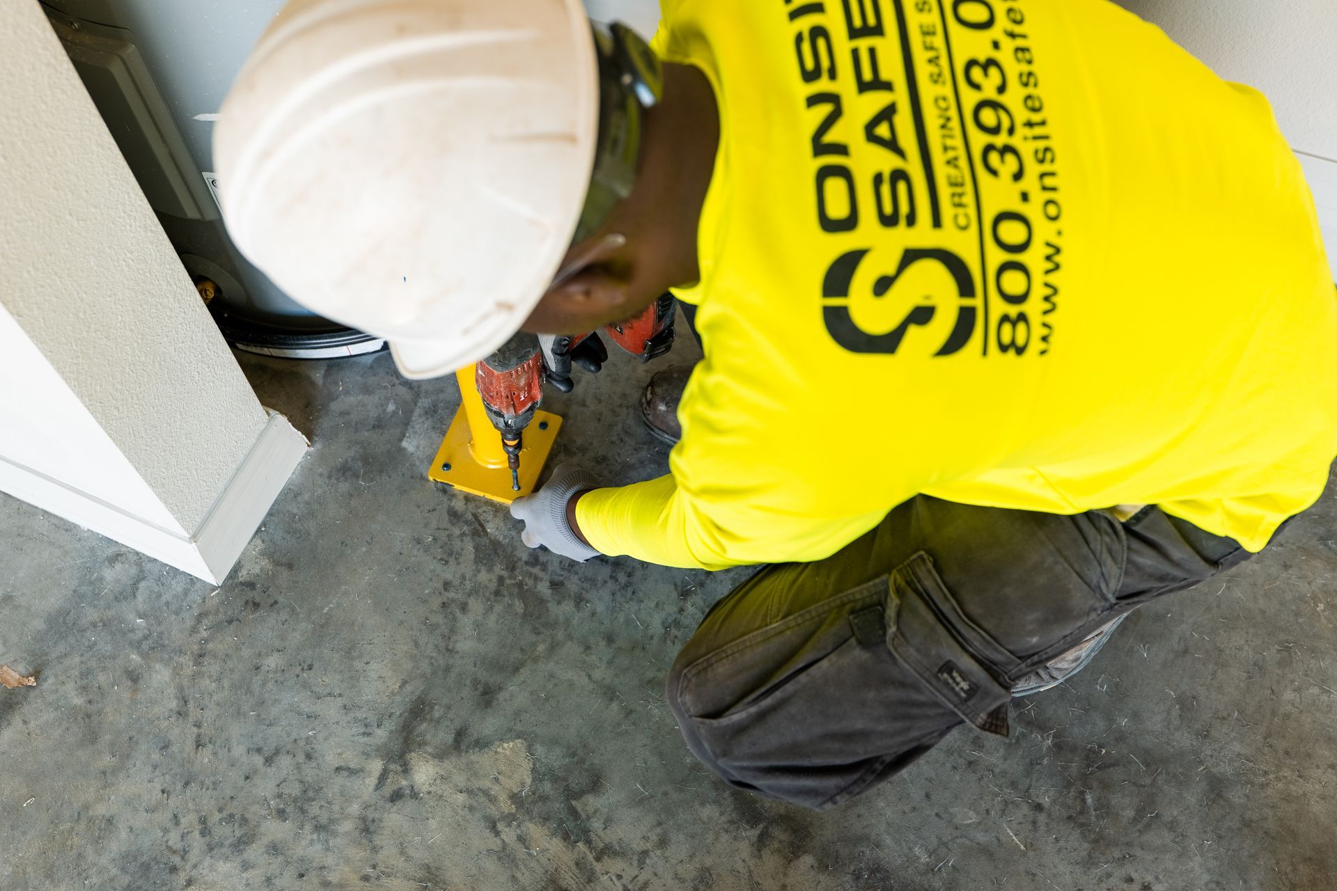 Worker in a yellow safety shirt kneels on a concrete floor, handling equipment near a wall.
