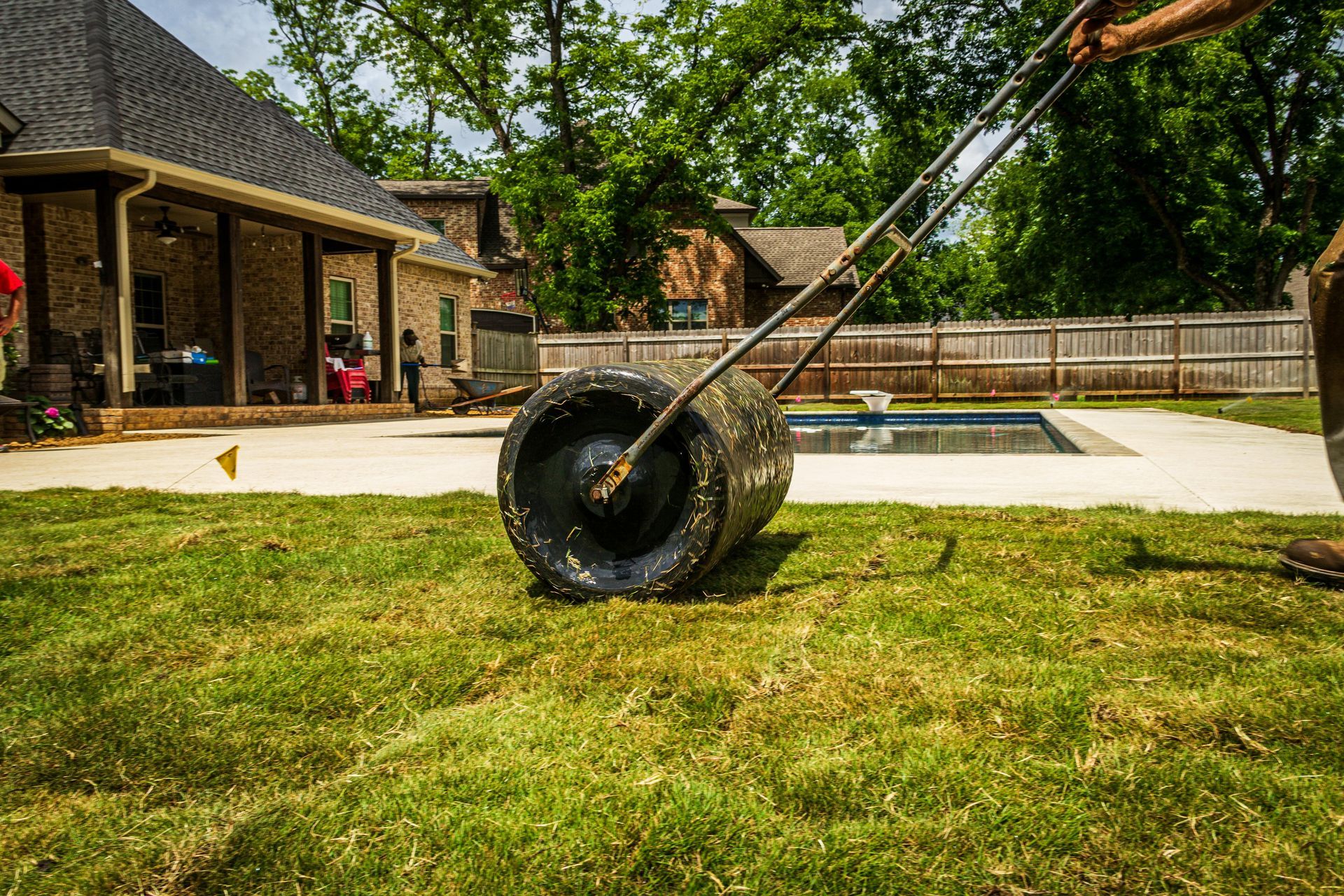Person rolling a lawn roller on grass near a house and swimming pool.