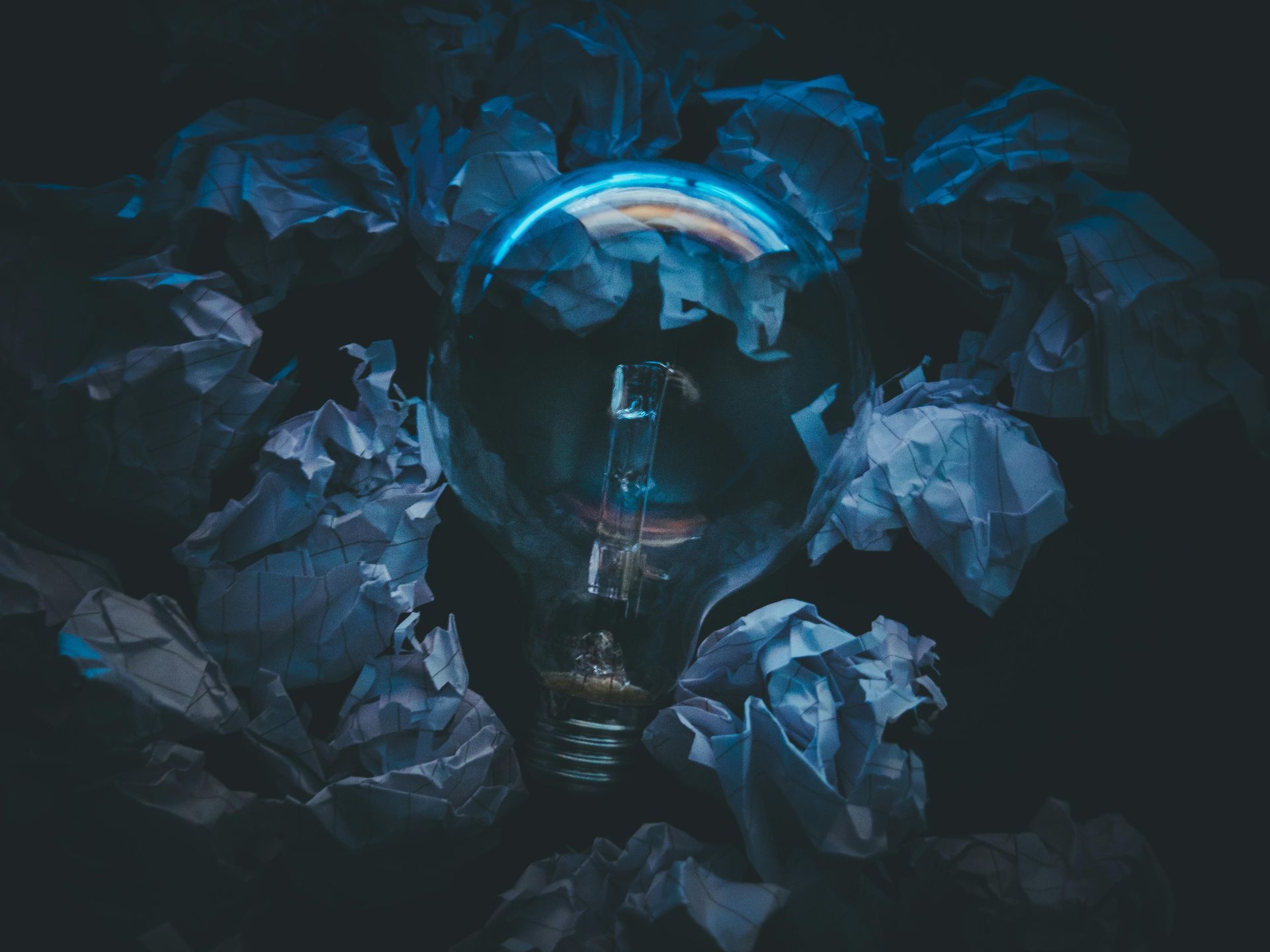 Glowing lightbulb surrounded by crumpled paper, illuminated by blue light in a dark setting.