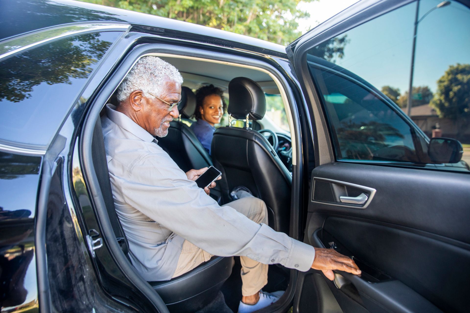 Man exiting a black car, holding a phone. Young woman sits in the driver's seat. Daytime