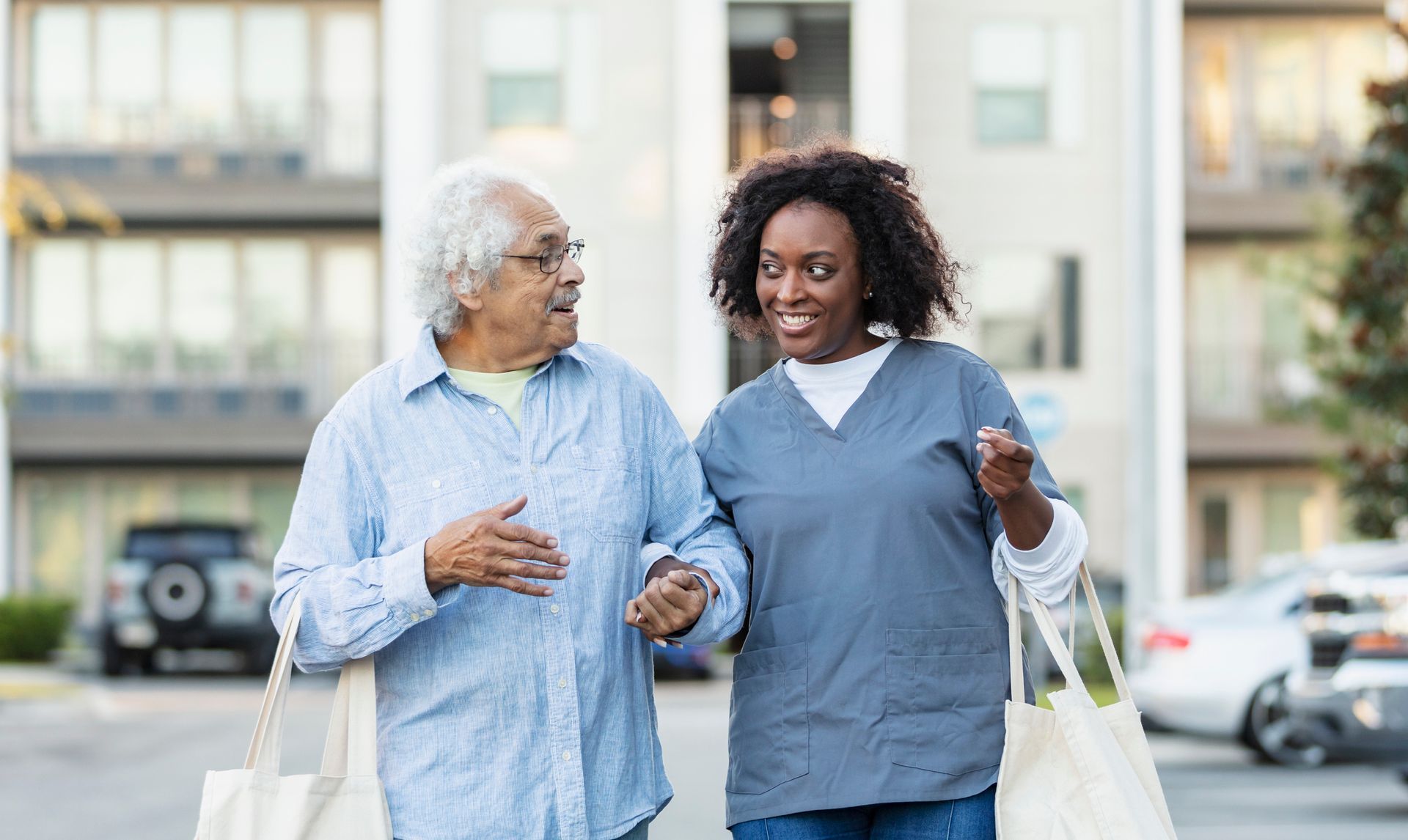 Older man and caregiver walking, carrying bags, outside apartment building.