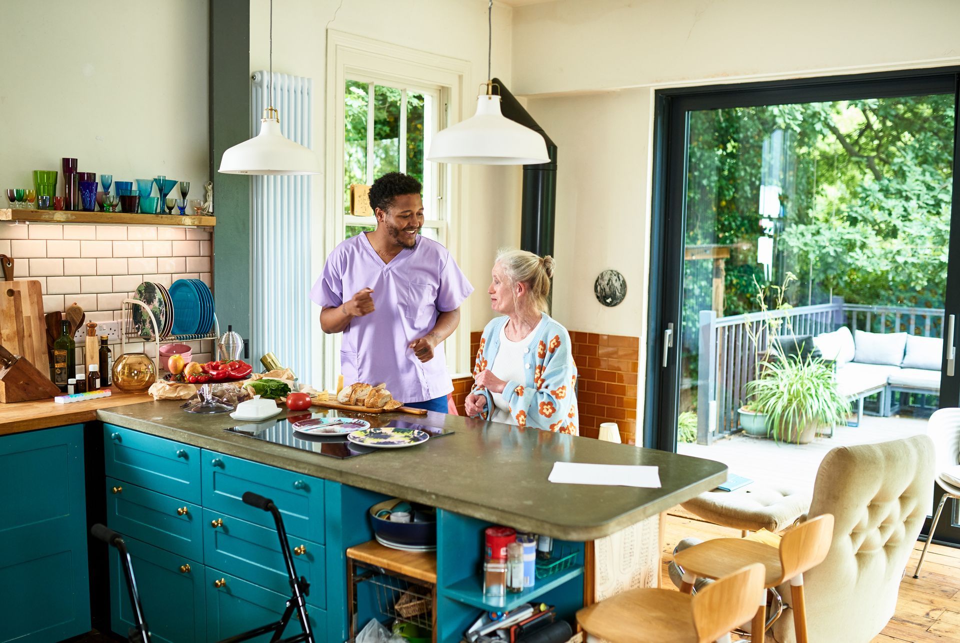 Caregiver and senior woman in kitchen; woman smiles, speaking. Sunny interior with food and deck view.