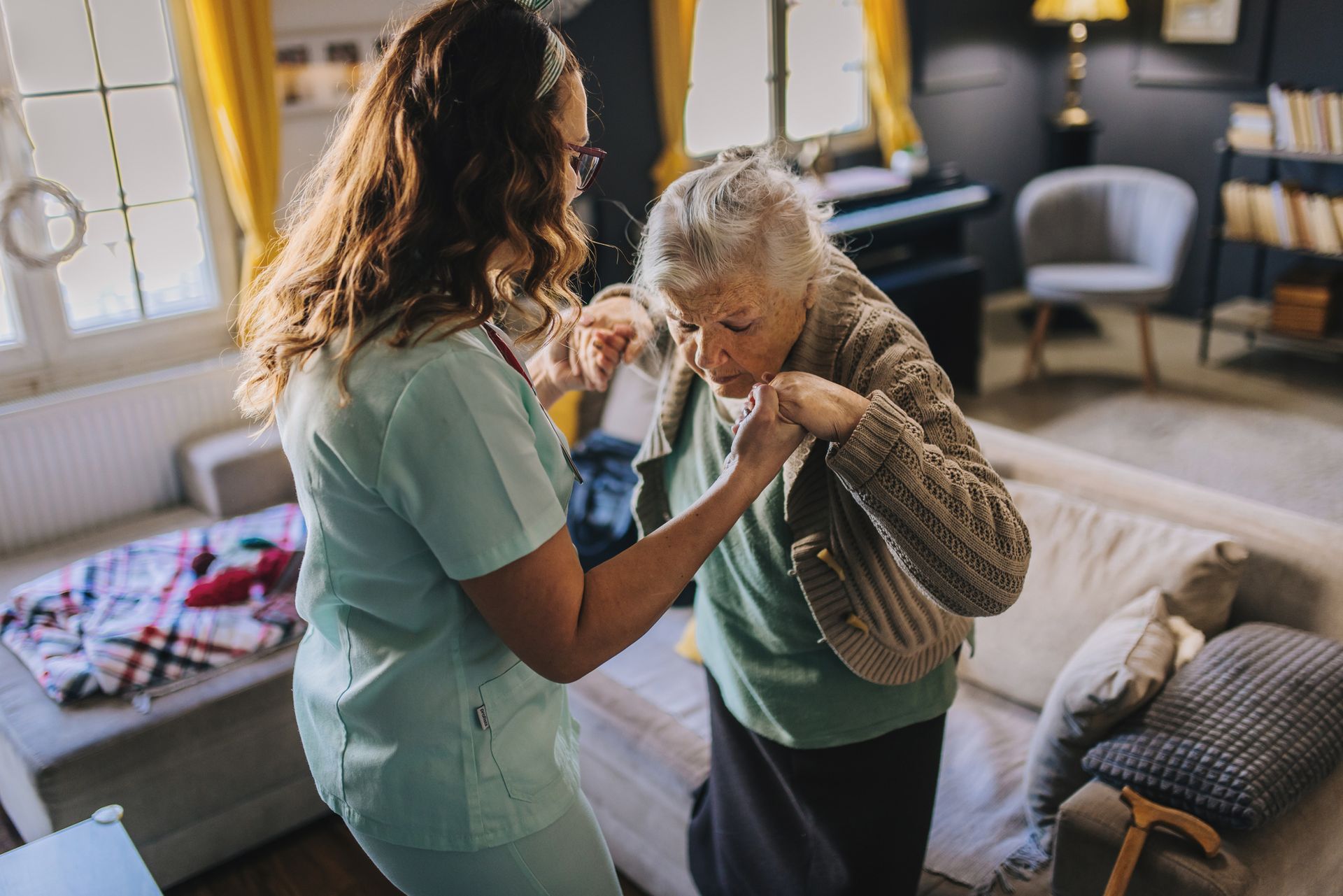 A caregiver helps an elderly woman stand up in a living room.